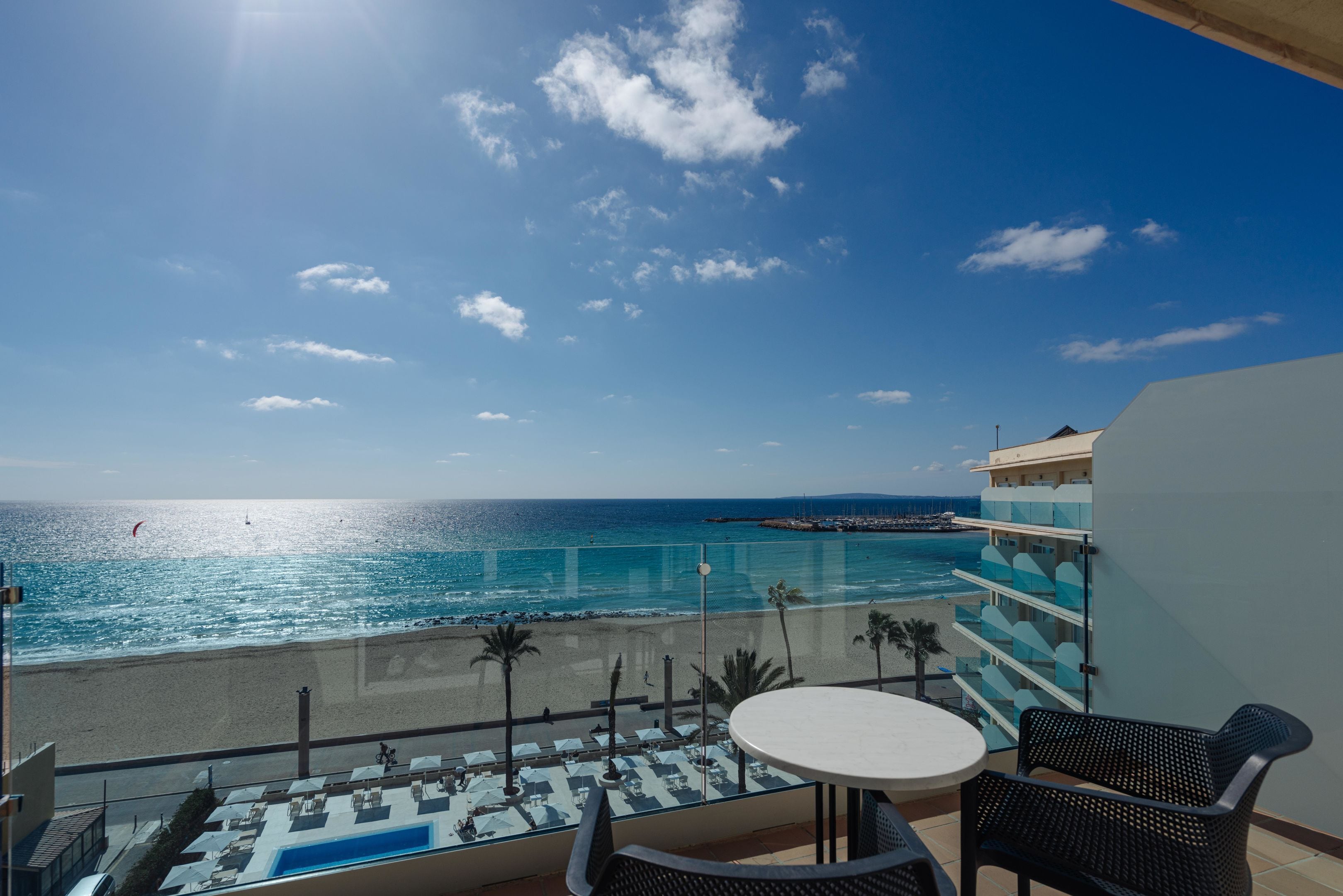 A balcony with a view of the beach and ocean, a pool area, and a table and chairs.