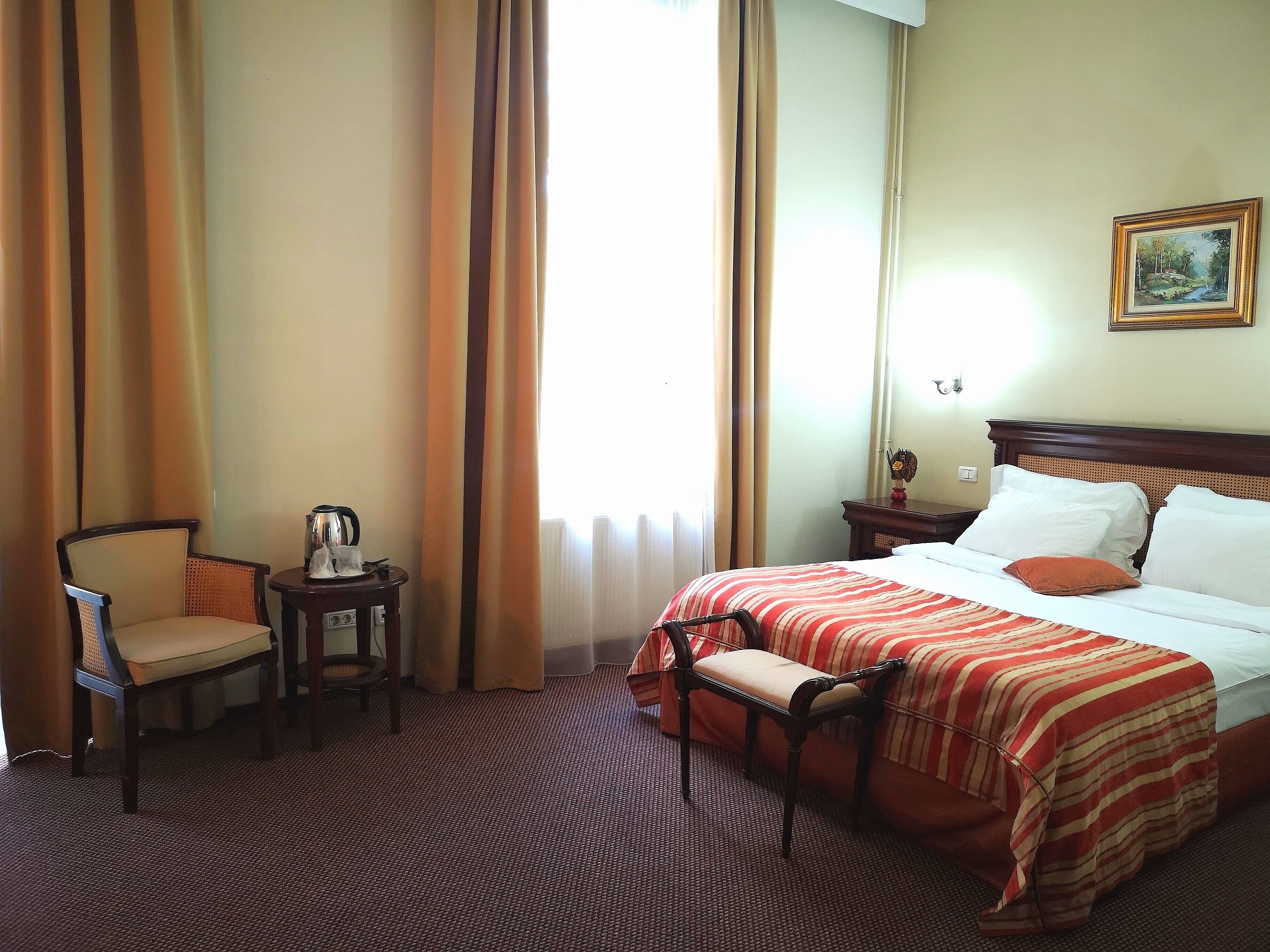 A hotel room with a bed, a chair, a small table with a kettle, and a painting on the wall.