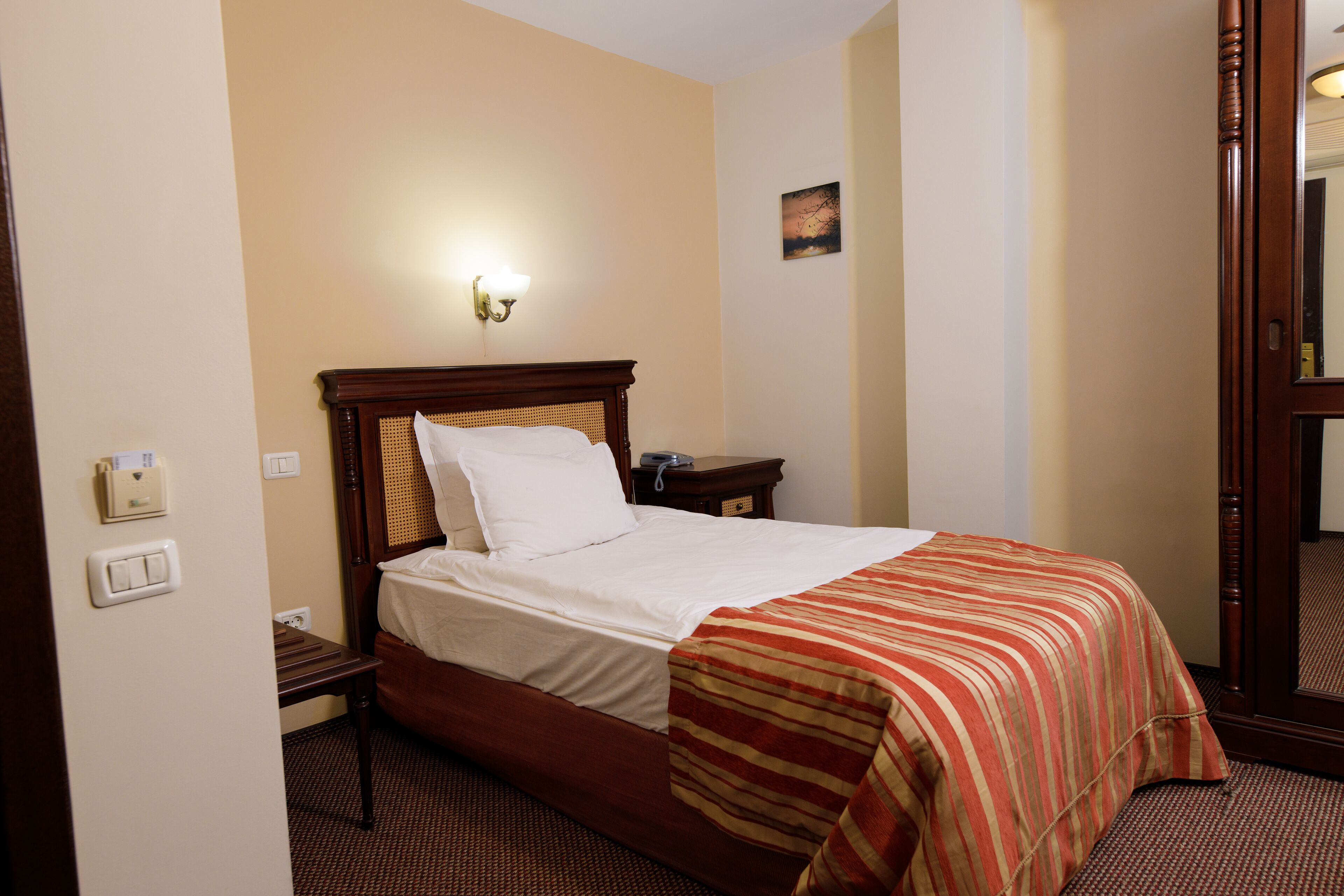 A hotel room with a wooden bed, a bedside table, a wall-mounted light, and a painting on the wall.