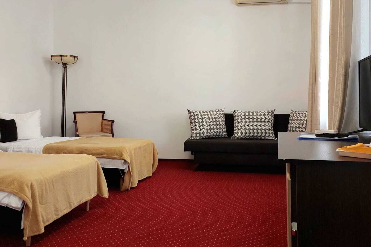 A hotel room with two beds, a sofa, a desk with a computer, and an air conditioning unit on the wall.