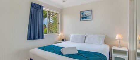Une chambre à coucher bien rangée, avec un lit, deux tables de chevet dotées de lampes, un ventilateur de plafond, une fenêtre avec des rideaux et un tableau encadré accroché au mur.