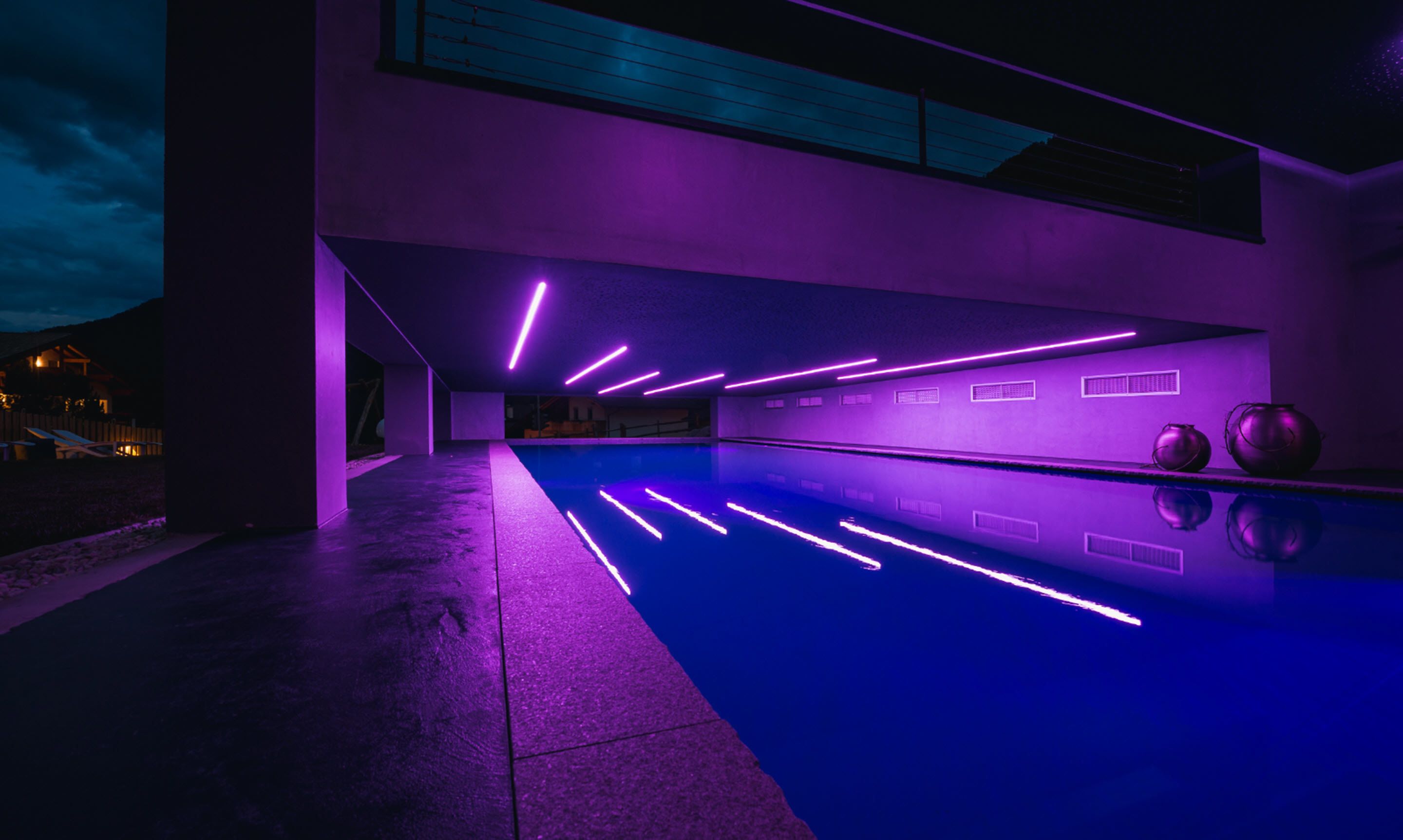 A modern indoor swimming pool with purple lighting, a concrete floor, and a view of the outside at night.