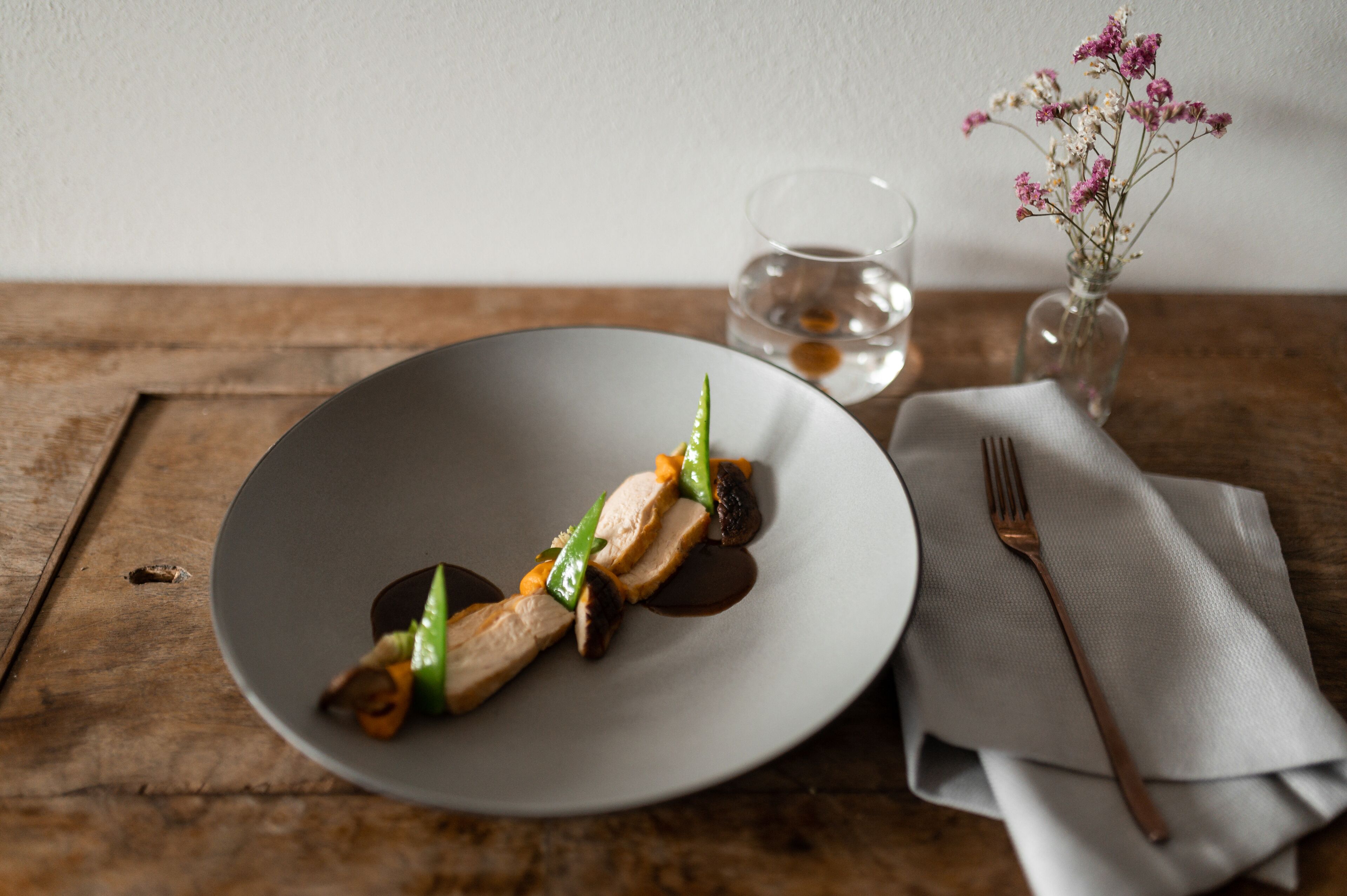 Un plato de comida sobre una mesa de madera, con un vaso de agua y un jarrón de flores al fondo.
