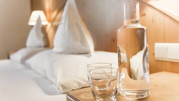 Une chambre d’hôtel avec une tête de lit en bois, un lit recouvert de draps blancs, une table de chevet sur laquelle se trouvent une bouteille transparente et un verre, et une lampe en arrière-plan.