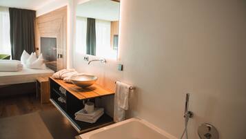 Una habitación de hotel moderna con una cama grande, un tocador de madera con lavabo y un espejo amplio, además de un área de ducha independiente.