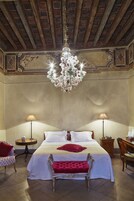 Habitación de hotel clásica con una cama grande, un banco tapizado en rojo, dos mesitas de noche con lámparas y un candelabro.
