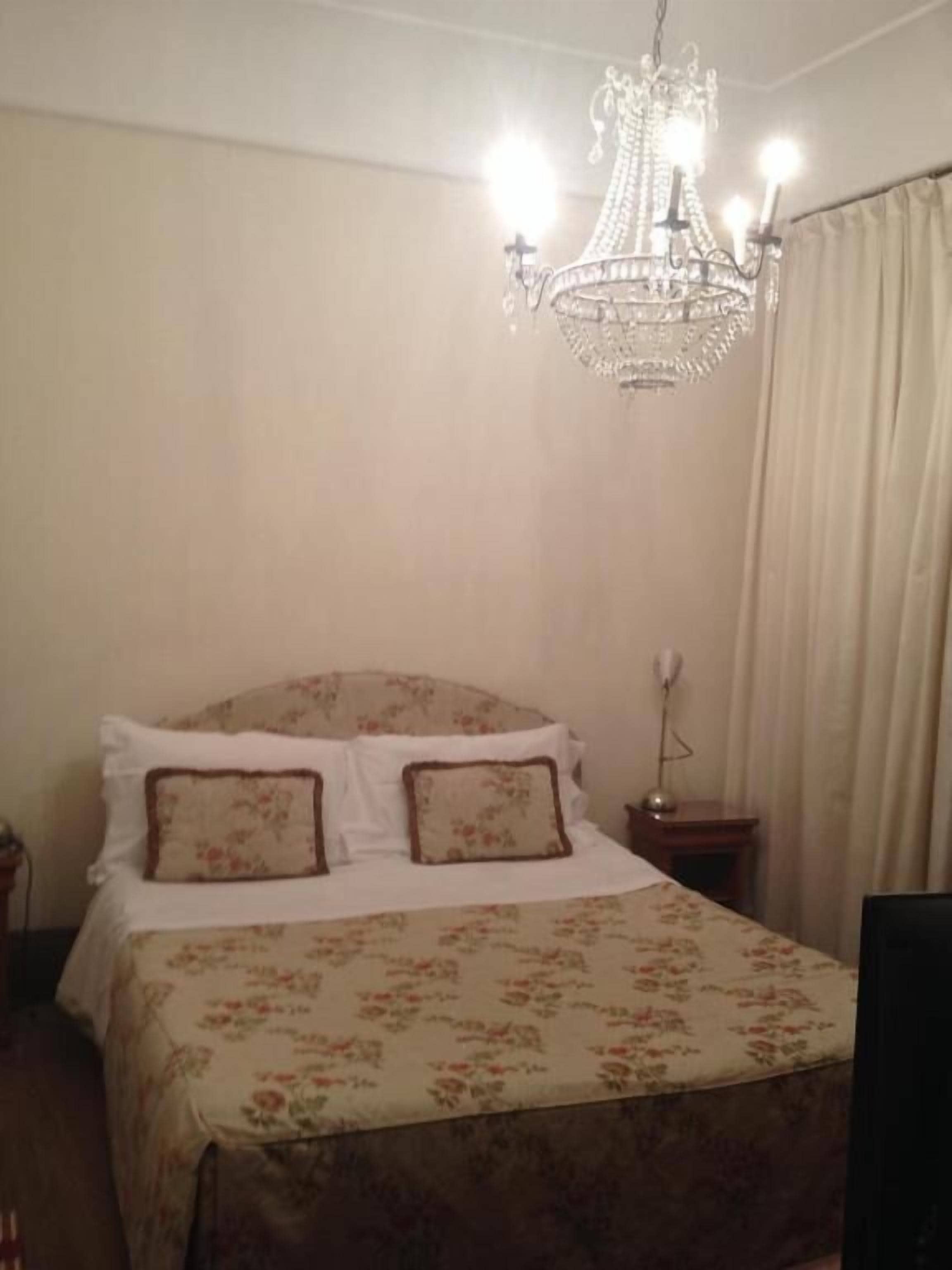 A bedroom with a bed, floral bedspread, two pillows, a chandelier, a nightstand, and a curtain.