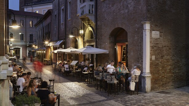 An outdoor cafe with tables and chairs, a cobblestone street, and buildings with brick facades. - Albergo Cappello (Ravenna)