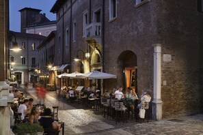 An outdoor cafe with tables and chairs, a cobblestone street, and buildings with brick facades. - Albergo Cappello (Ravenna)