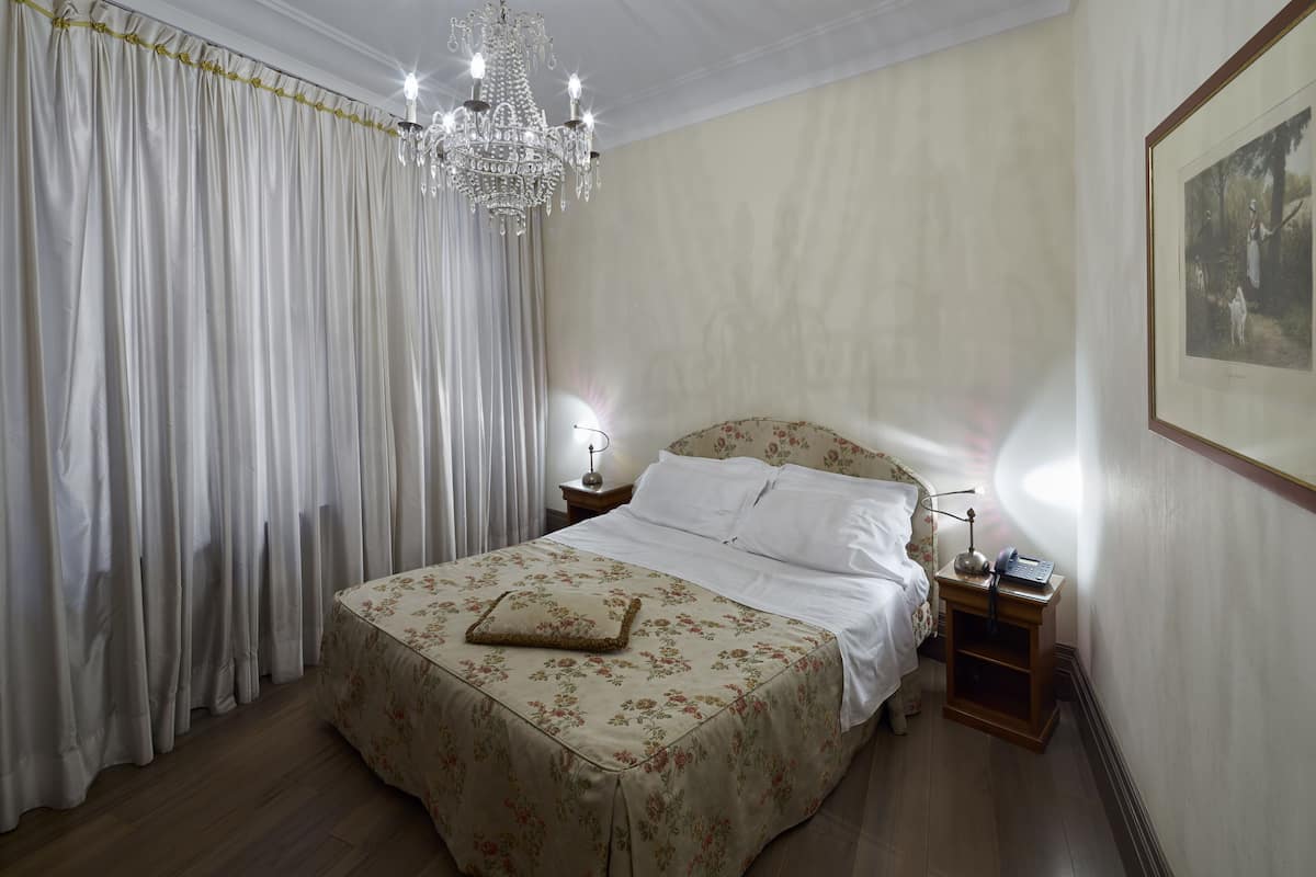 A bedroom with a large bed, a chandelier, a nightstand with a lamp, and a framed picture on the wall.