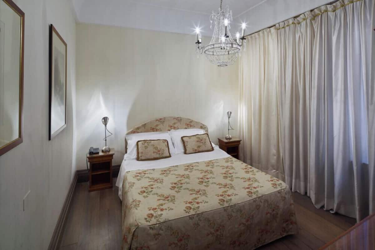 A bedroom with a large bed, a chandelier, a nightstand, and a window with curtains.