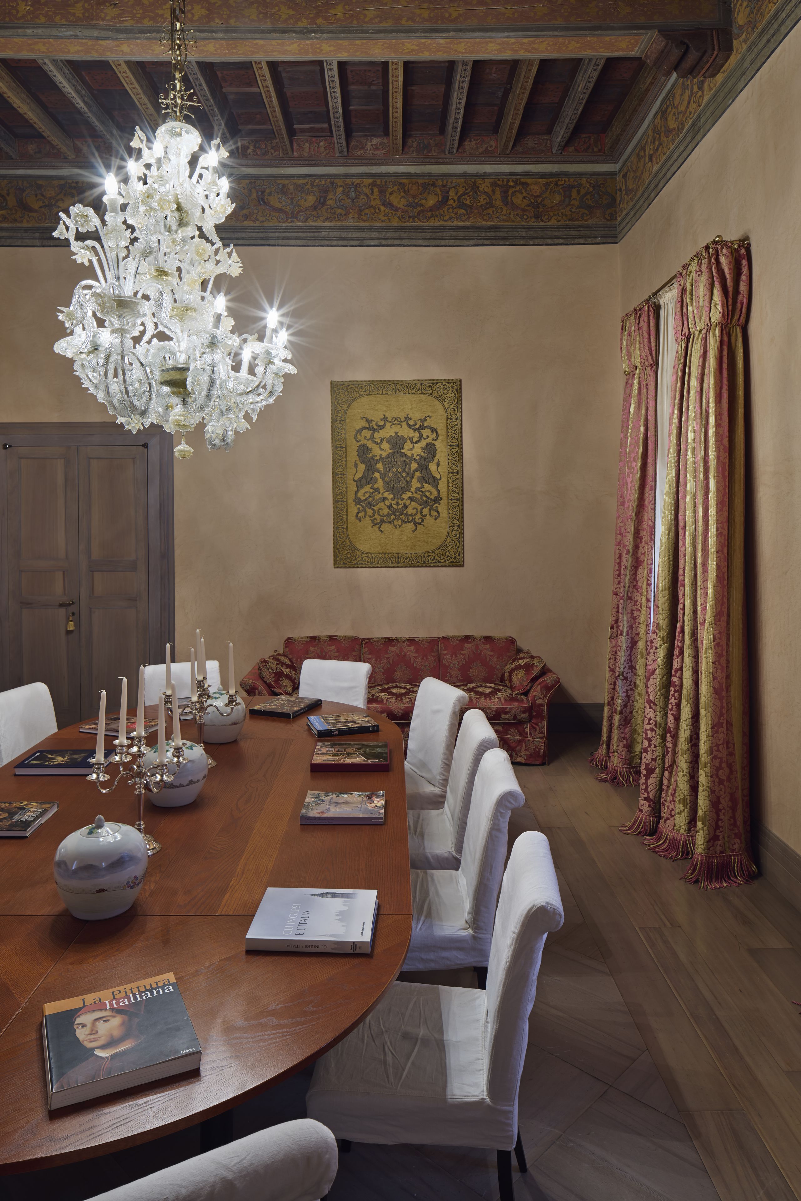 Un comedor con una mesa de madera, sillas blancas, una lámpara de araña y un sofá rojo.