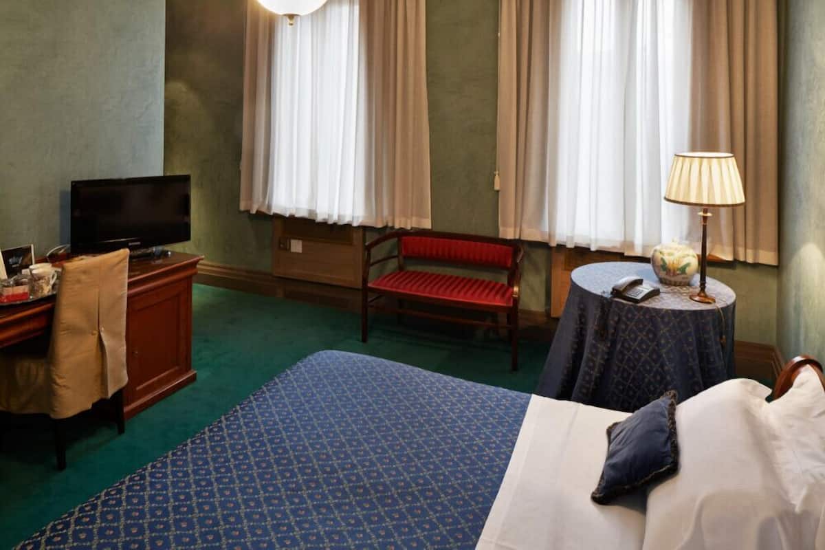 A hotel room with a bed, a desk with a television, a red chair, a round table with a lamp, and a window with curtains.
