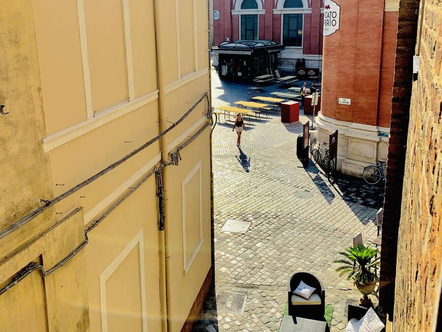 Una calle estrecha con pavimento adoquinado, mobiliario exterior con mesas y sillas, y un edificio con un letrero que dice "CATO PERIO".