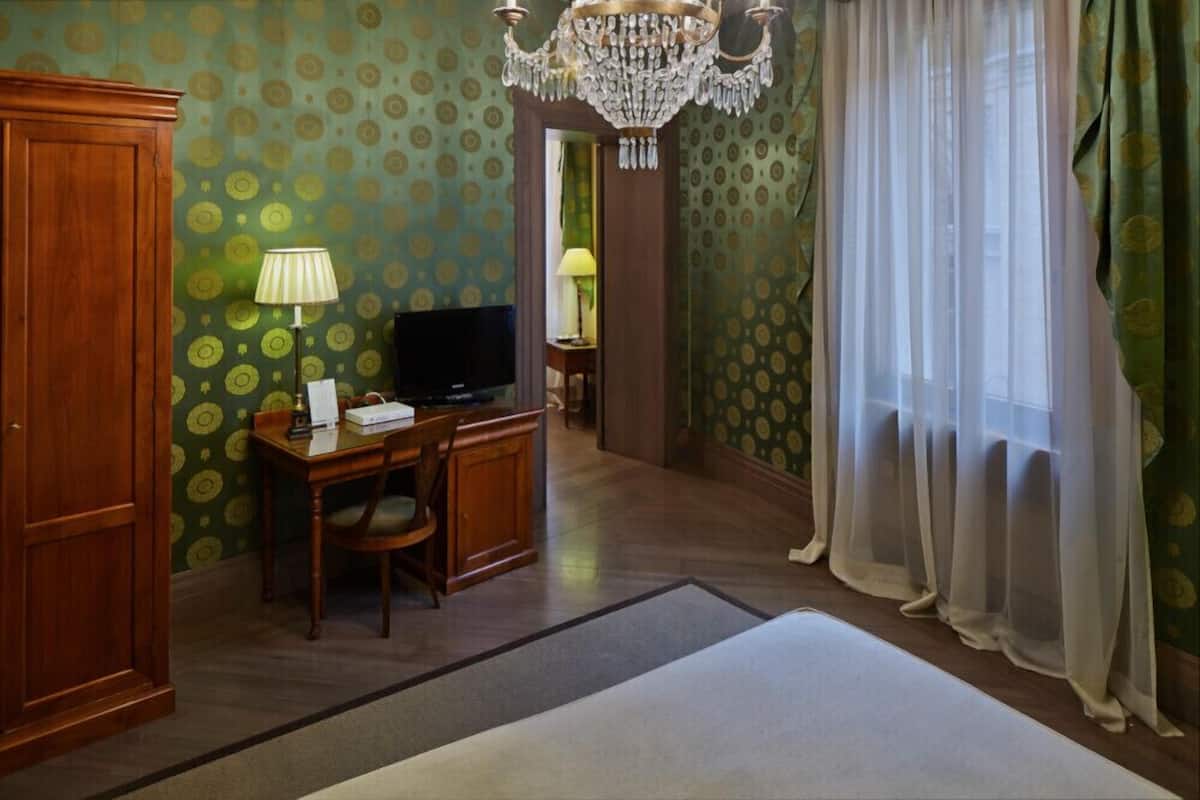 A hotel room with a wooden desk, a television, a chandelier, and a large window with sheer curtains.