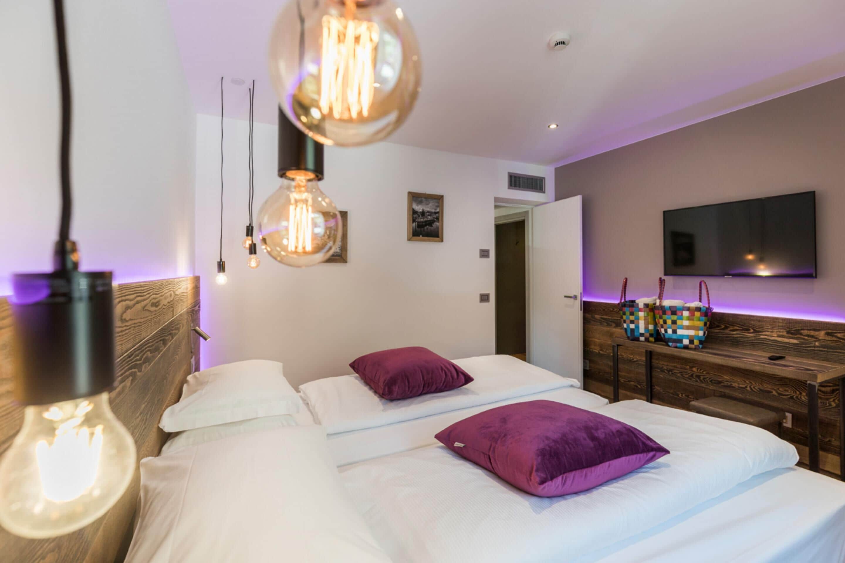 Una habitación de hotel moderna con dos camas, almohadas moradas, un cabecero de madera, un televisor de pantalla plana y lámparas colgantes.