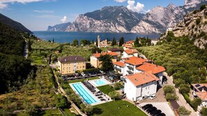 A coastal hotel with a swimming pool, multiple buildings, and a mountainous backdrop. - Aktivhotel Santalucia (Nago-Torbole)