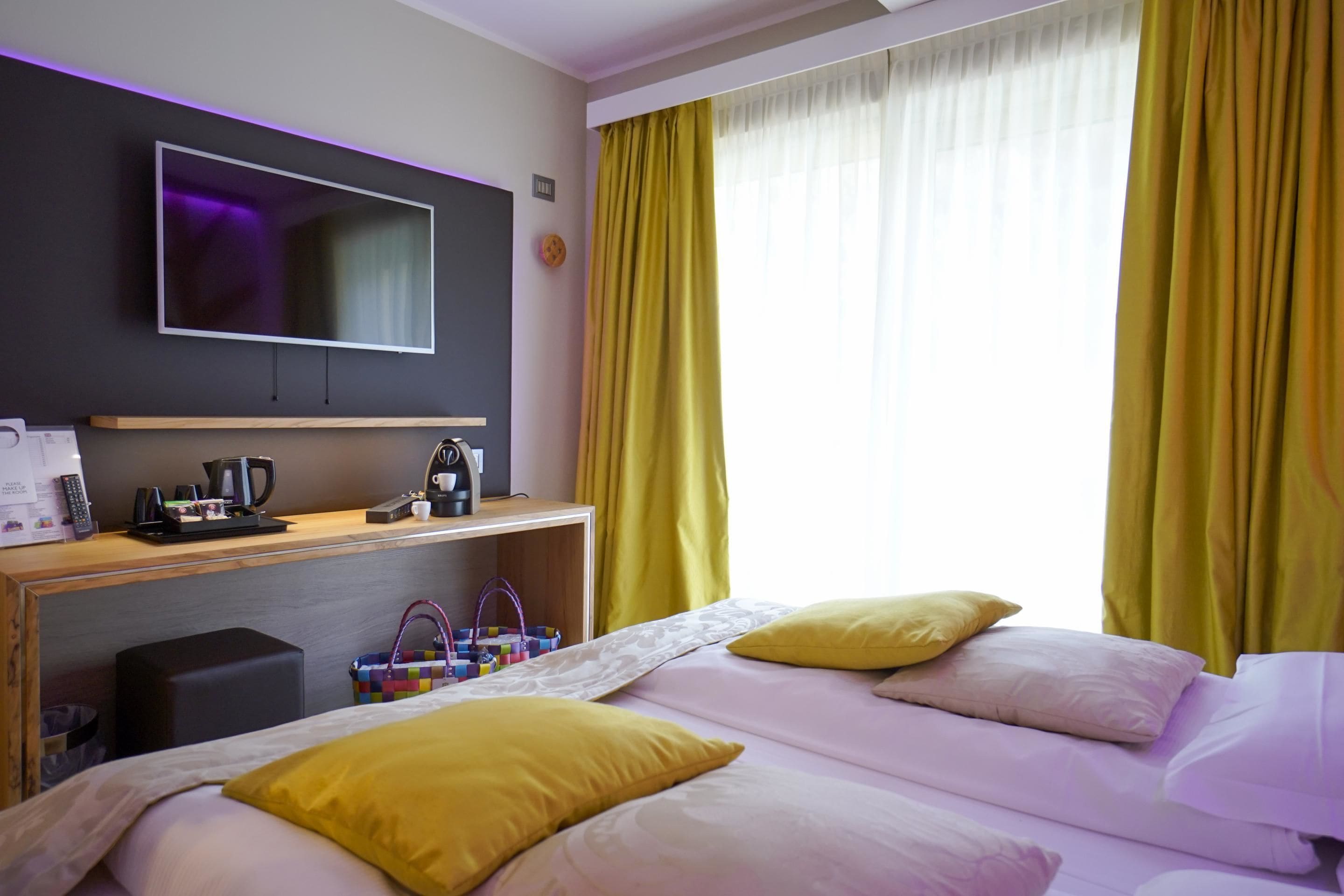Une chambre d’hôtel avec un lit, un téléviseur fixé au mur, un bureau avec une machine à café et des rideaux jaunes.
