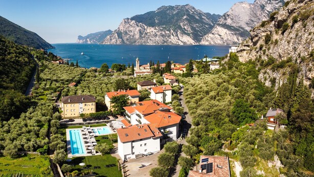 A coastal village with a swimming pool, terraced houses, and a mountain backdrop. - Aktivhotel Santalucia (Nago-Torbole)