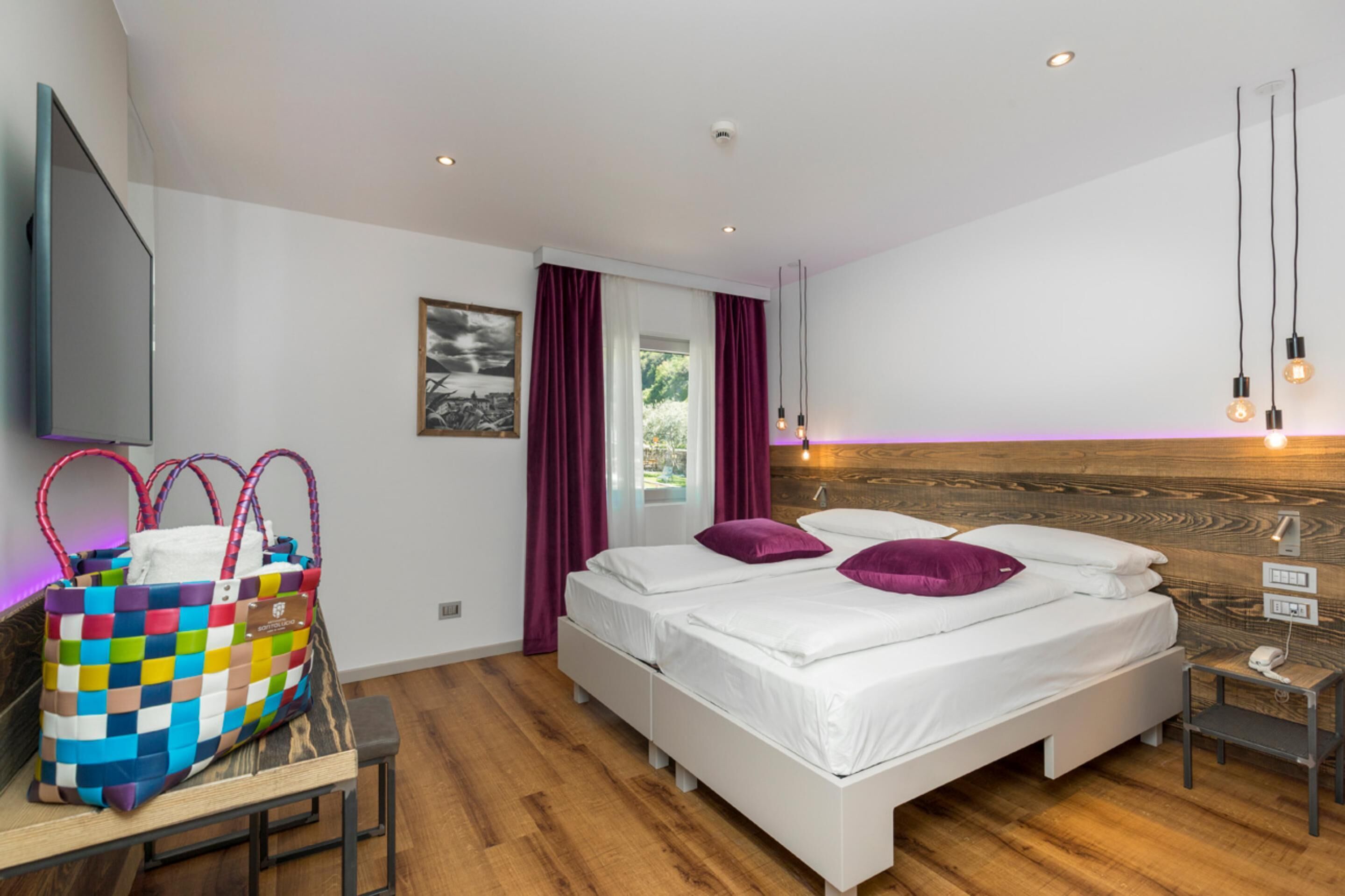 Une chambre d’hôtel moderne avec une tête de lit en bois, deux lits avec du linge de lit blanc et des oreillers violets, un téléviseur fixé au mur, un sac coloré posé sur un banc et une fenêtre avec des rideaux.