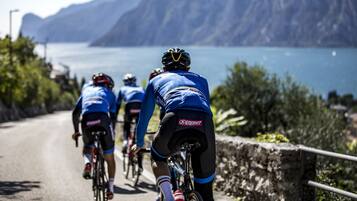 Cyclists on a road with a stone wall and greenery on one side, and a body of water and mountains in the background.
