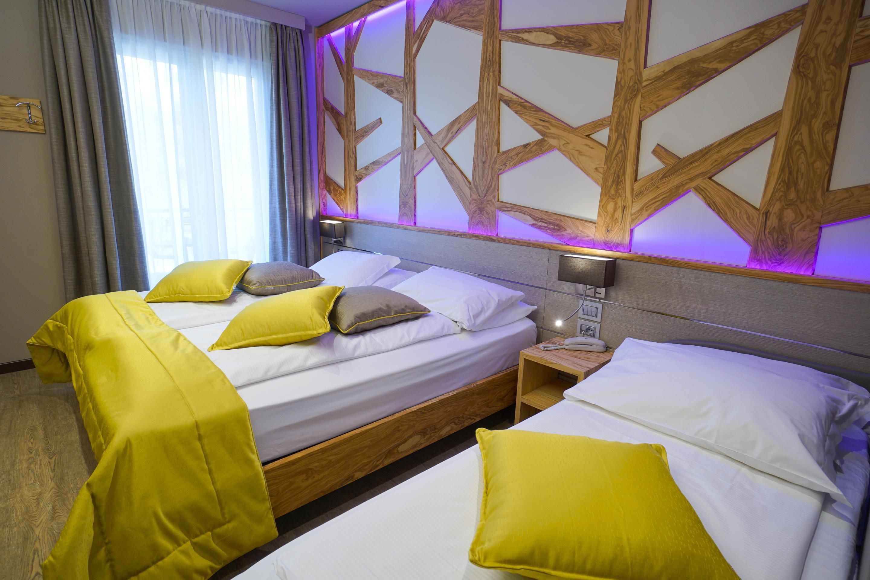 Une chambre d’hôtel avec deux lits, chacun agrémenté de draps blancs et d’oreillers jaunes. On y trouve une tête de lit en bois ornée d’un motif géométrique et éclairée d’une lumière violette. Une table de chevet avec une lampe est visible à côté des lits.