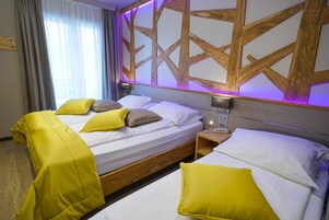 Habitación de hotel con dos camas, cada una con ropa de cama blanca y almohadas amarillas. Se aprecia un cabecero de madera con diseño geométrico e iluminación morada. Junto a las camas, un mesita de noche con una lámpara.