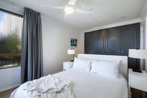 Una cama bien tendida con sábanas blancas, un ventilador de techo, una mesita de noche con lámpara y una ventana con cortinas.