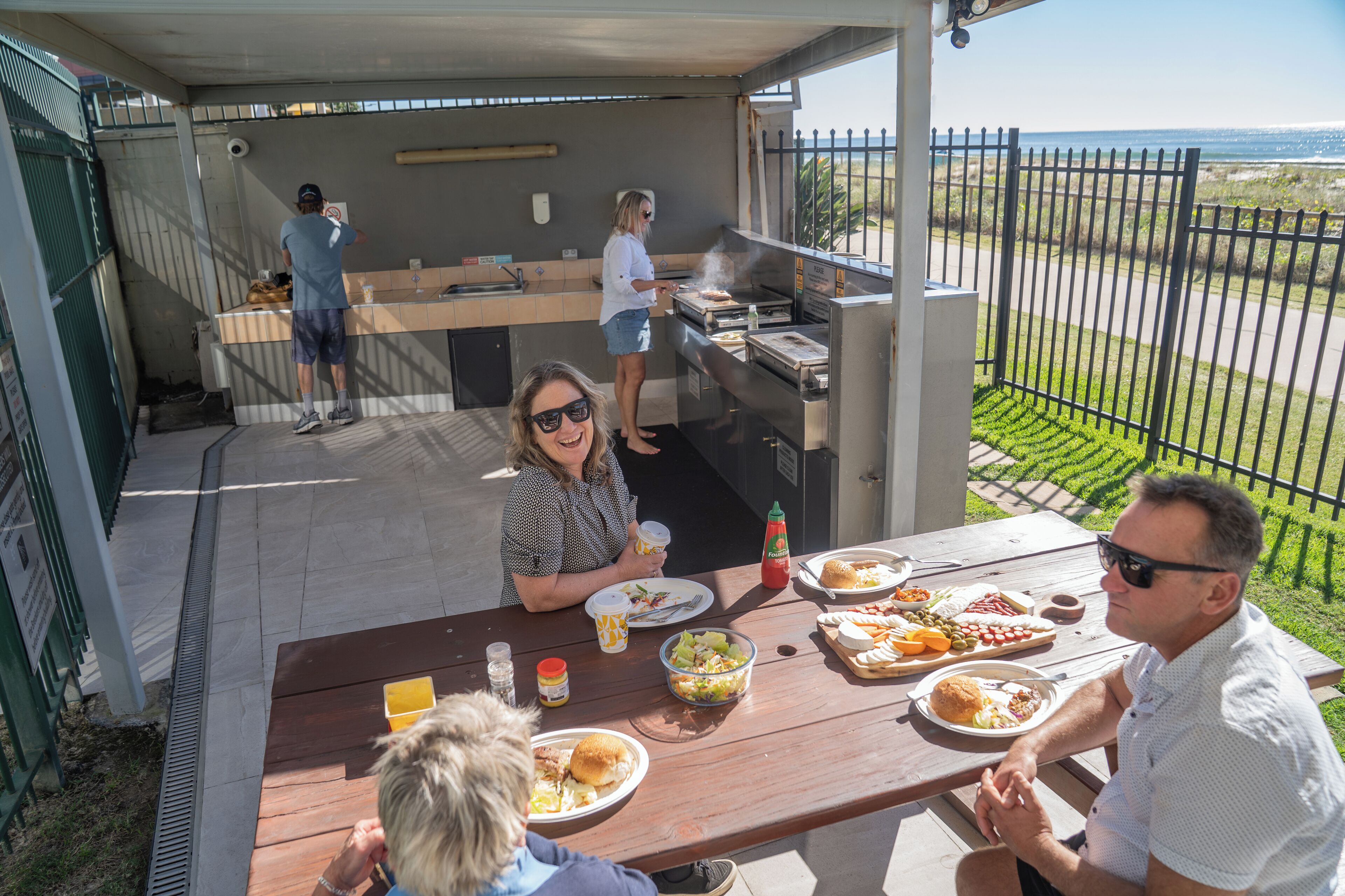 An outdoor dining area with a grill station, a picnic table with food, and a view of the ocean.
