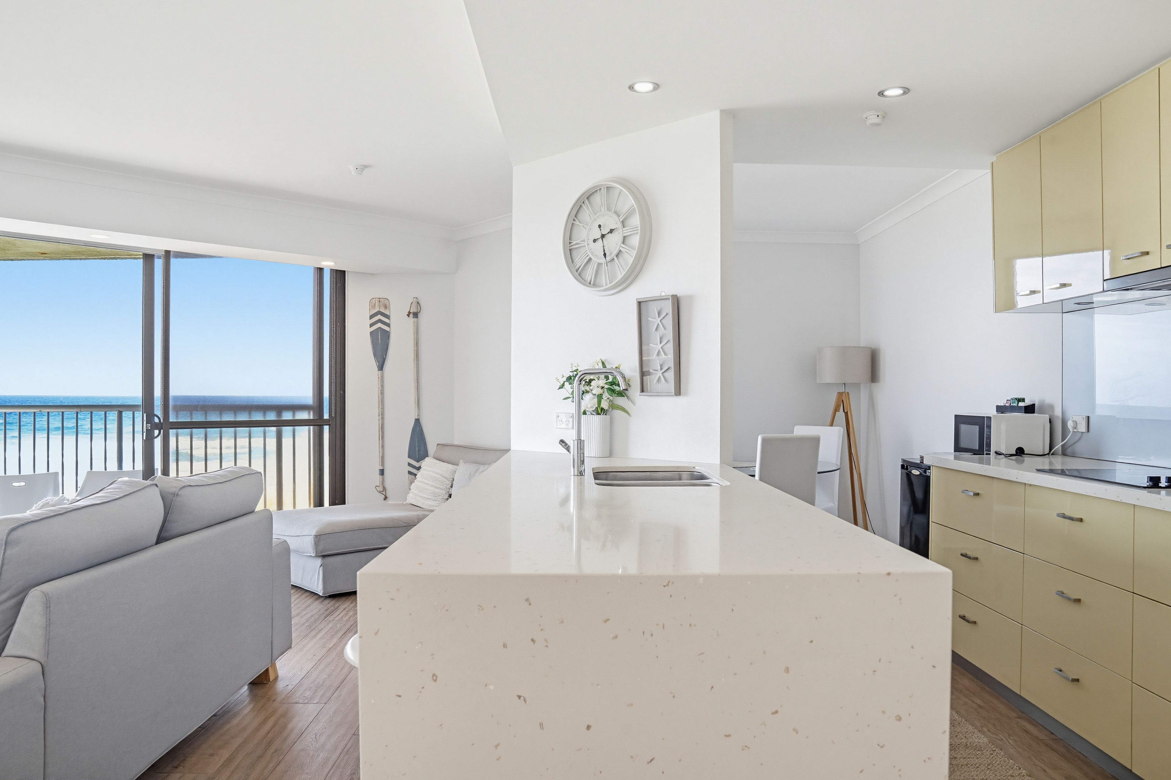 Une cuisine moderne avec un îlot central, un coin repas offrant une vue sur la mer et un salon avec un canapé et une grande fenêtre.