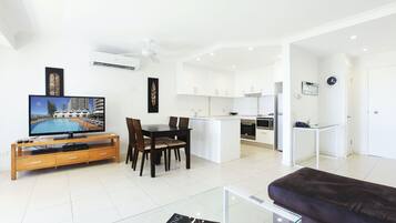 A modern living room with a TV, dining table, and sofa. There is a kitchen area with white cabinets and a small island.