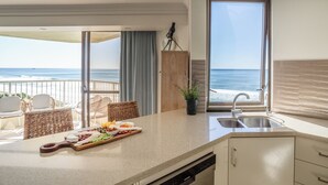 A kitchen with a view of the ocean, a dining area with wicker chairs, and a buffet table with various foods. - The Breakers (Surfers Paradise)