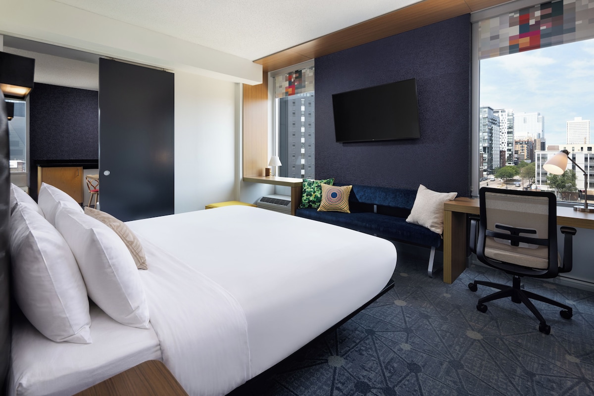 A hotel room with a bed, a desk, a chair, a TV, and a city view.