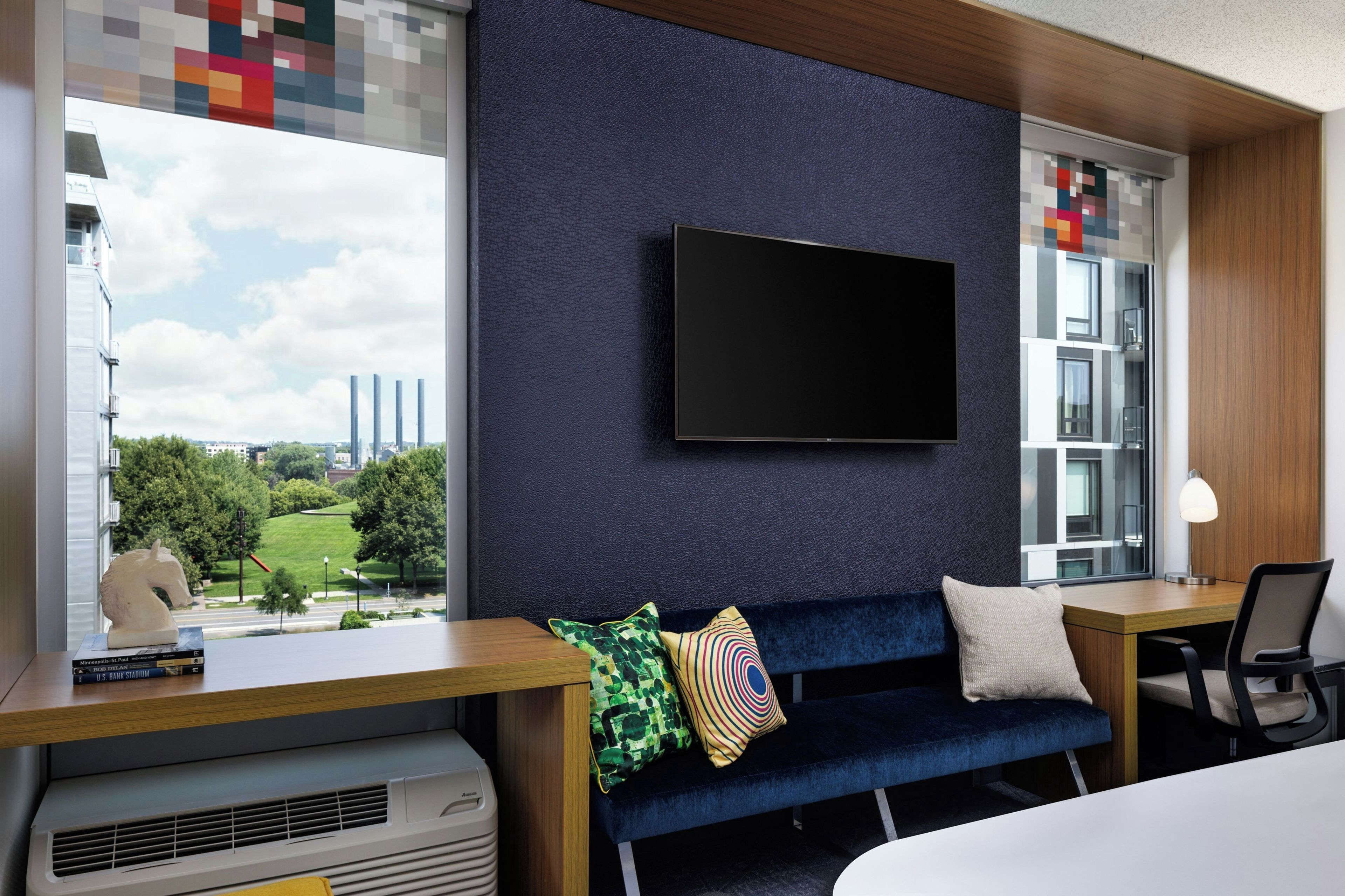 Una habitación de hotel moderna con un ventanal grande, un televisor de pantalla plana en la pared, un sofá azul, un escritorio con una lámpara y vistas a un paisaje urbano.
