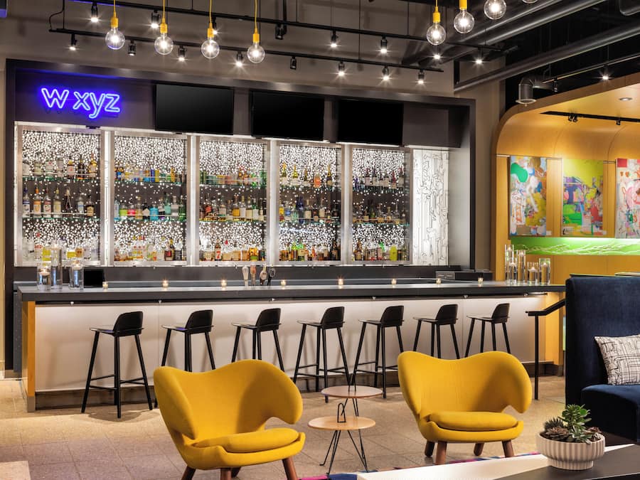 A bar area with a neon sign, a bar counter with stools, and a seating area with yellow chairs.
