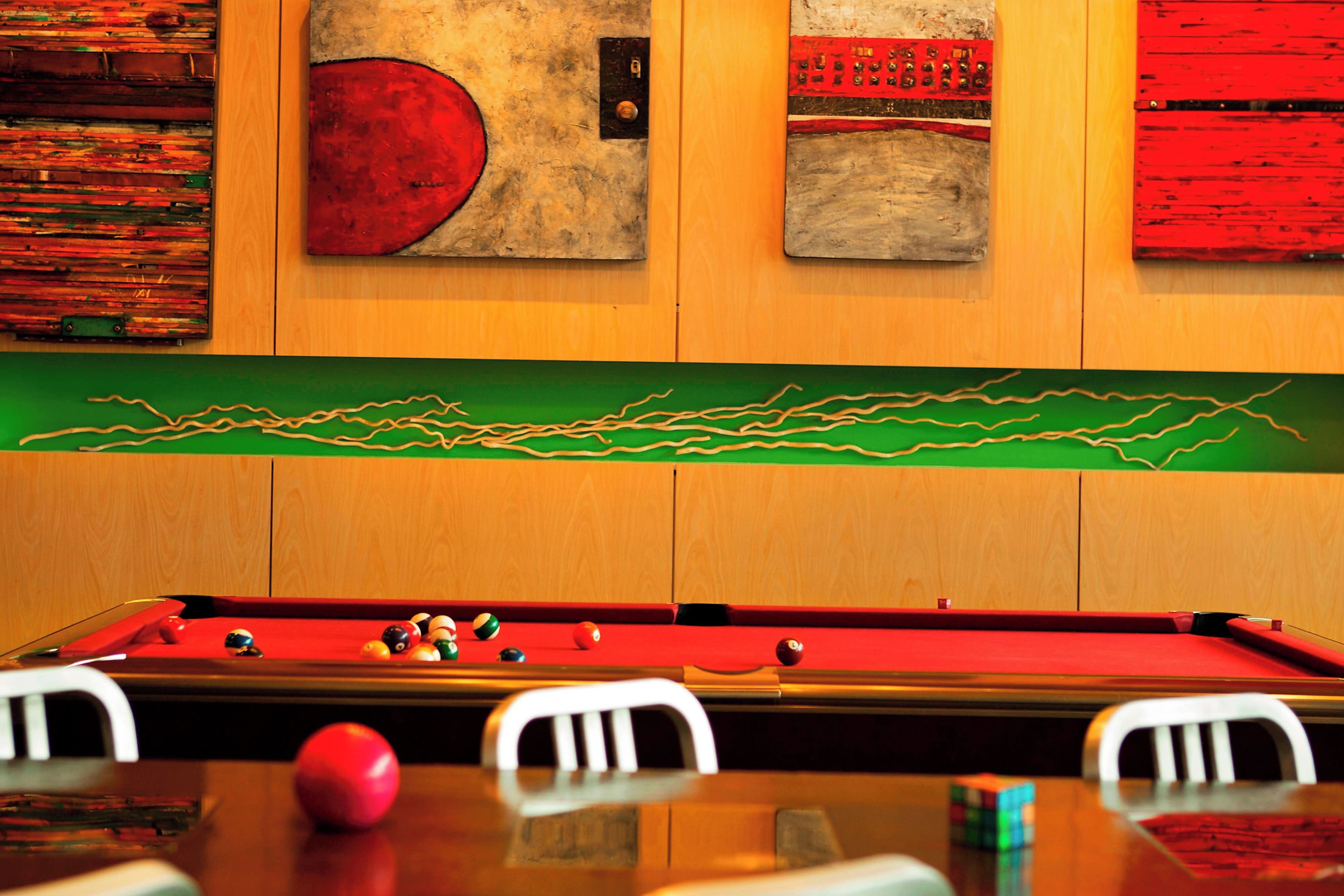 A pool table with balls and chairs, a red billiard cue, and a wall with abstract art and a green painted line.
