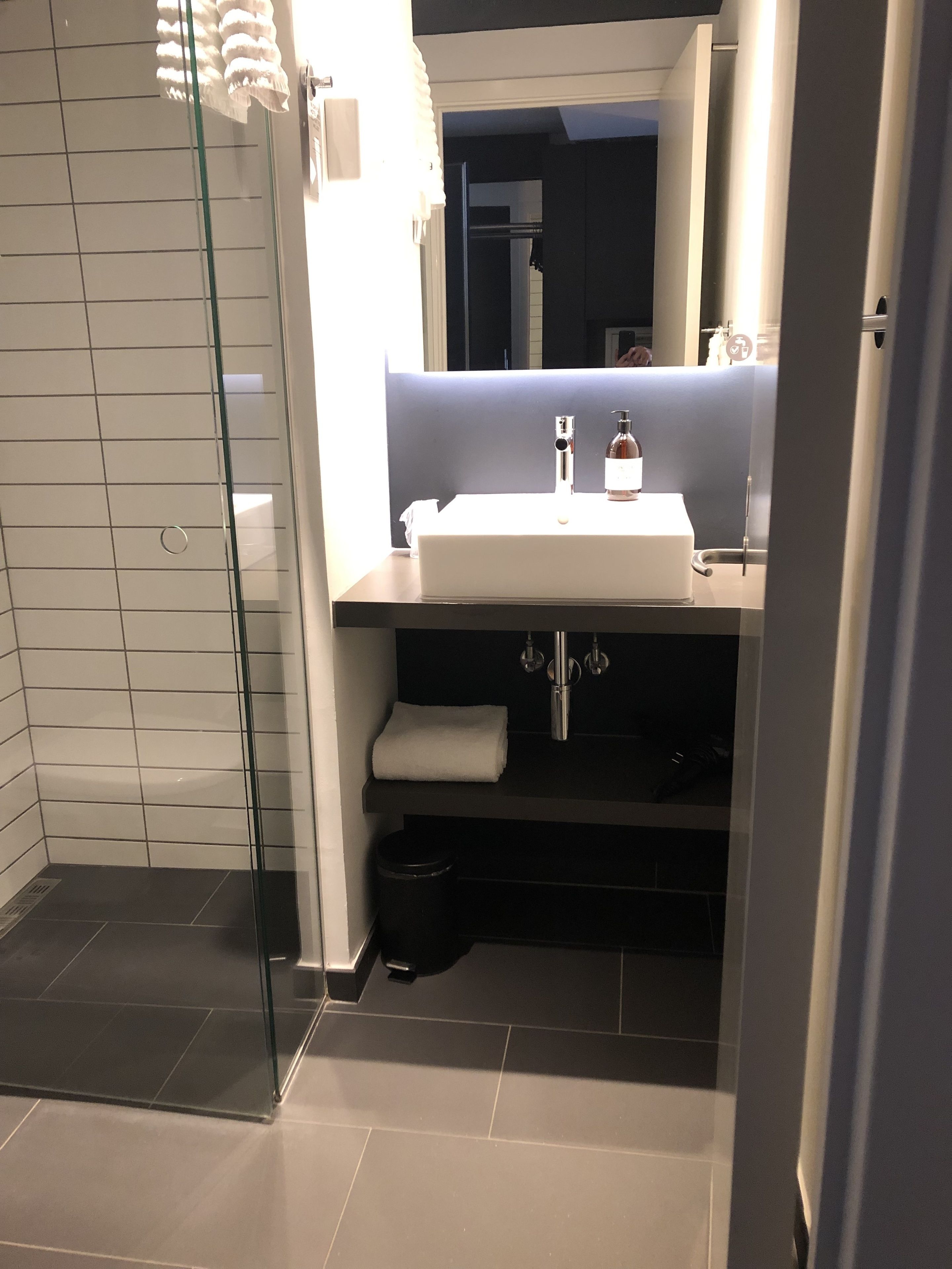 Ein modernes Badezimmer mit Duschabtrennung aus Glas, einem weißen Waschbecken mit Spiegel darüber und einer schwarzen Ablage darunter.