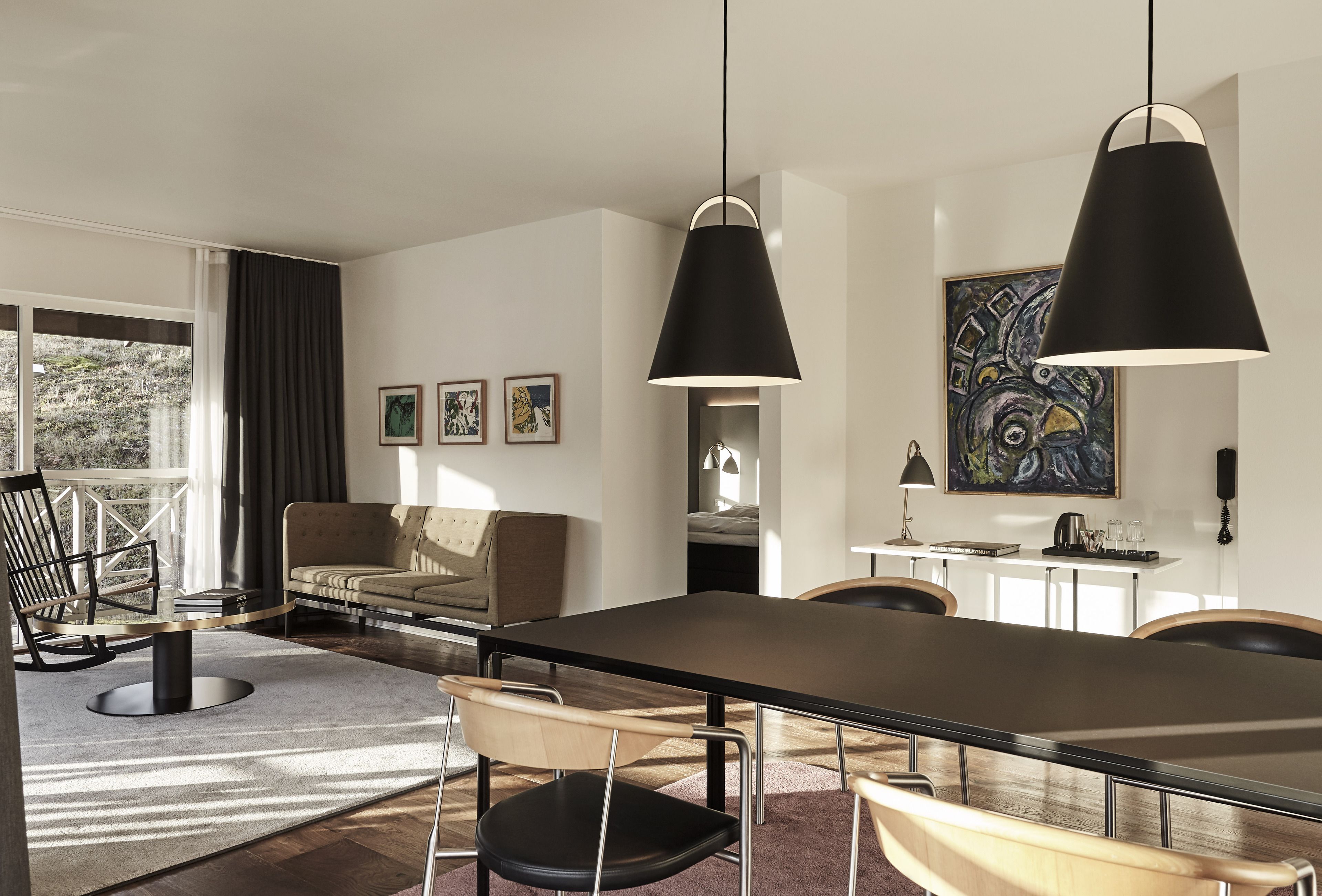 A modern dining area with a long table, chairs, and pendant lighting. There is a sofa and a chair in the background, and artwork on the walls.