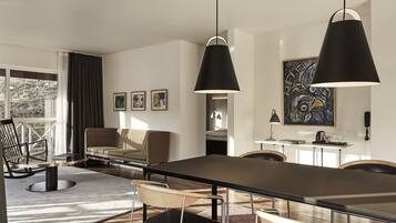 A modern dining area with a long table, chairs, and pendant lighting. There is a sofa and a chair in the background, and artwork on the walls.