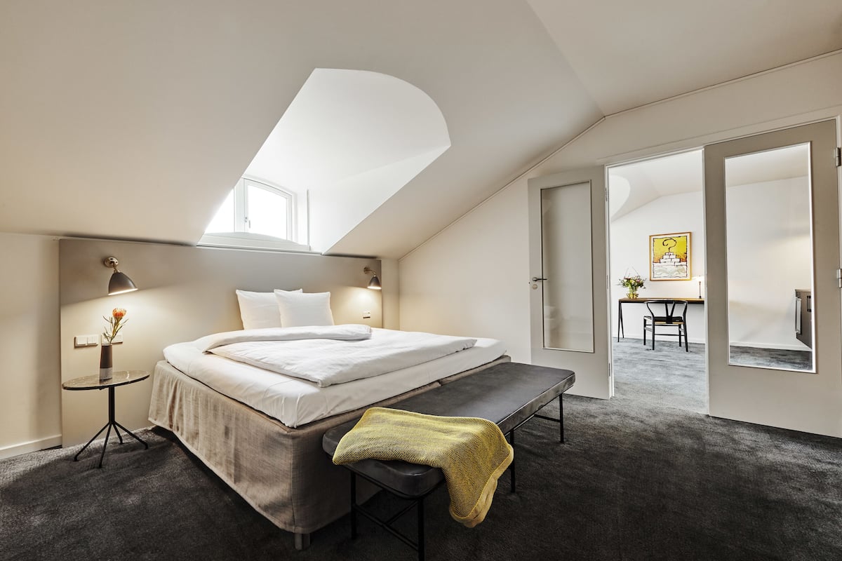 A modern bedroom with a skylight, a bed, a bench, and a small table with a lamp.