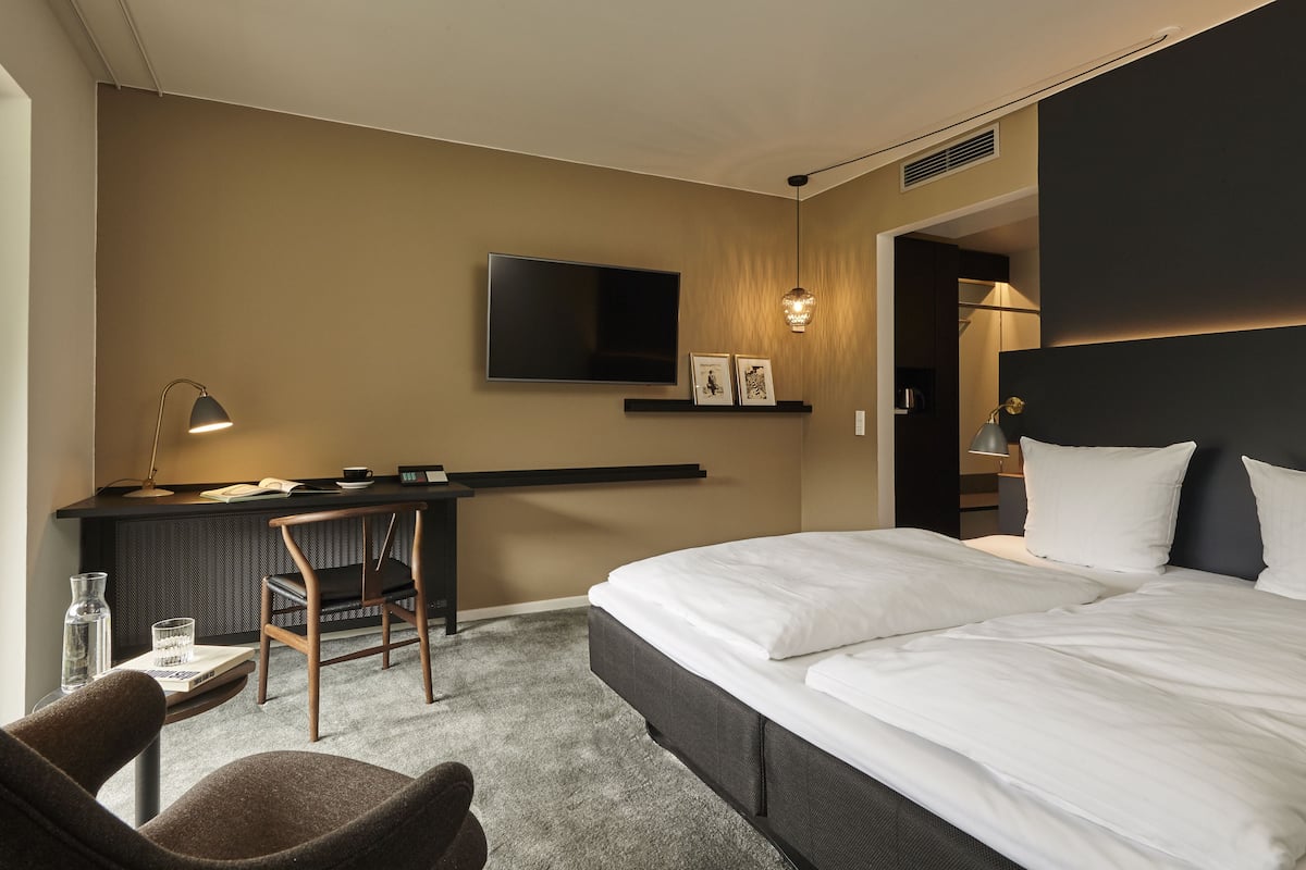 A hotel room with a bed, a desk with a chair, a TV, and a shelf with pictures.