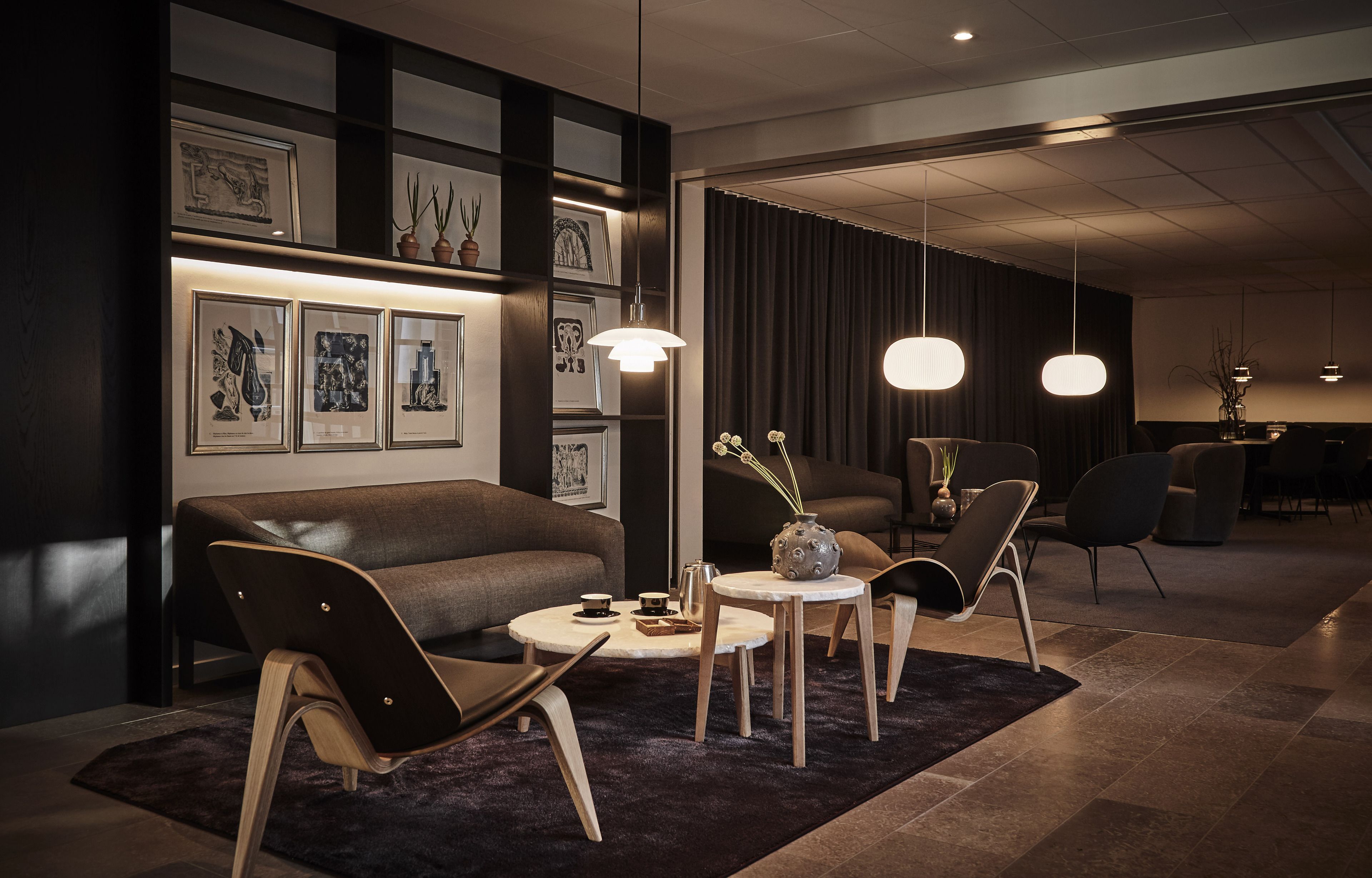 Een moderne hotellobby met een donker houten receptiebalie, een bank en een salontafel. Er hangen ingelijste kunstwerken aan de muur en hanglampen boven de zithoek.