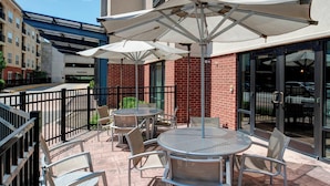 An outdoor patio with round tables and chairs, umbrellas, and a brick building in the background. - SpringHill Suites St. Louis Brentwood (Brentwood)