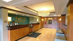 Ein Hotel-Rezeptionsbereich mit Holztresen, einer Uhr, gerahmten Bildern an der Wand und einer Sitzecke mit gemusterten Kissen.