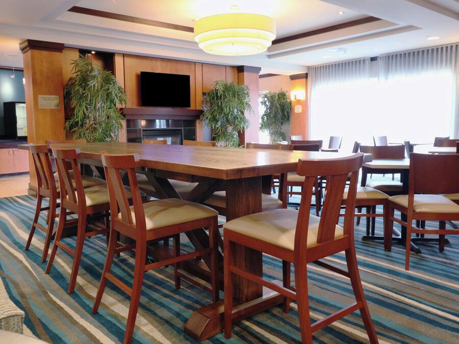 A hotel dining area with a large wooden table surrounded by chairs, a television, and potted plants.