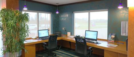 A conference room with two desks, computers, chairs, a large potted plant, and a window with a view of a parking lot.
