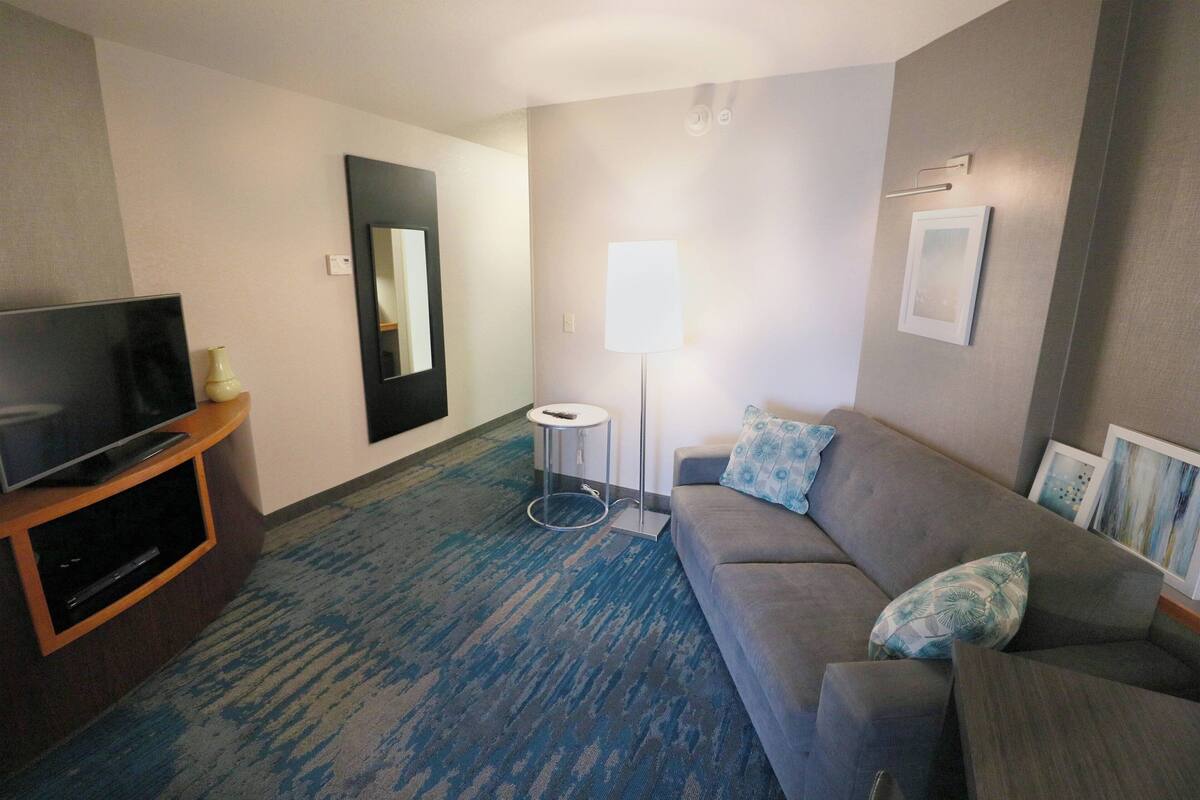 Une chambre d’hôtel avec un téléviseur à écran plat posé sur un meuble en bois, un canapé gris agrémenté de coussins à motifs, un lampadaire et un tableau encadré accroché au mur.