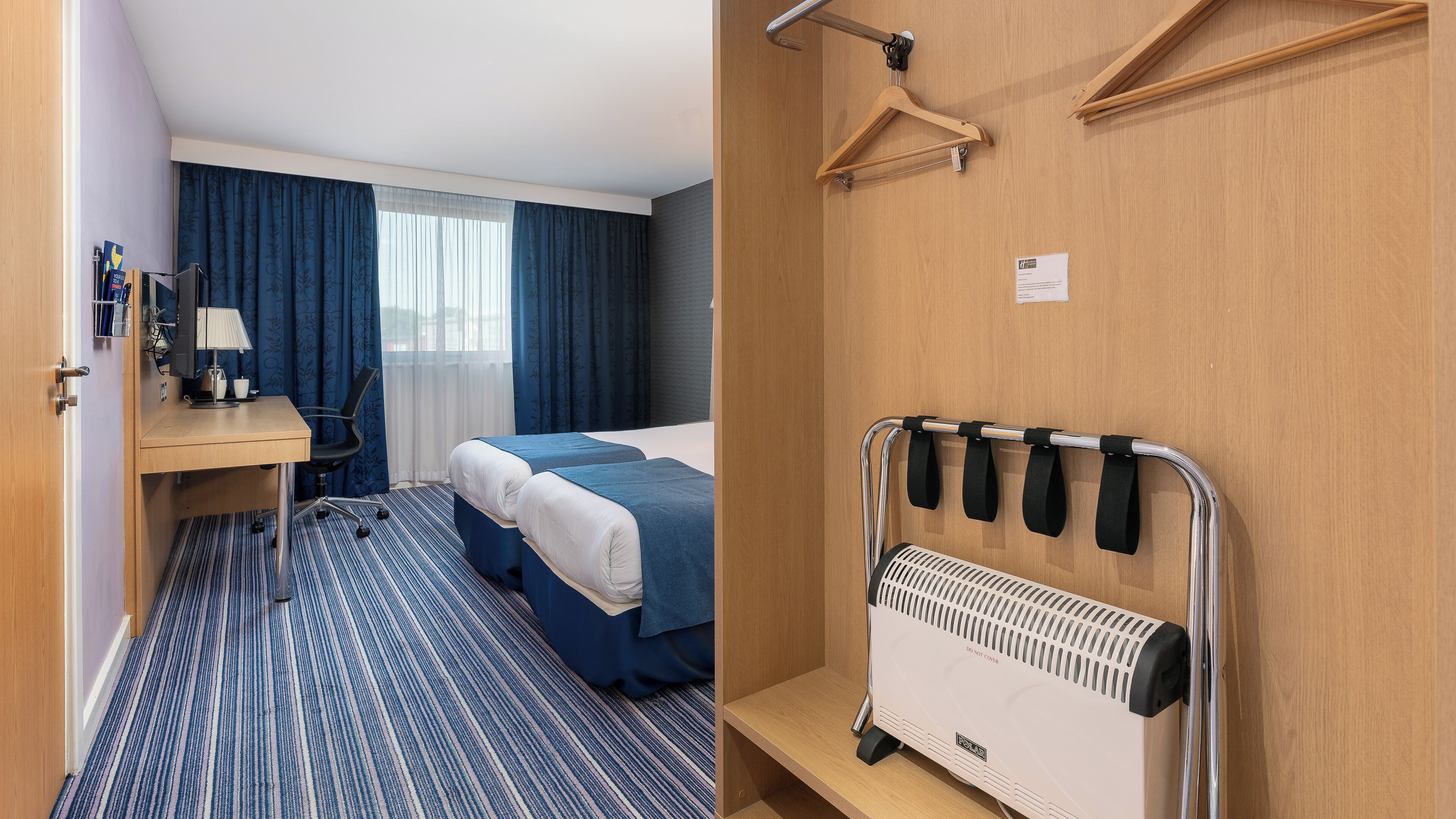 A hotel room with a bed, a desk, a chair, a radiator, and a wardrobe.