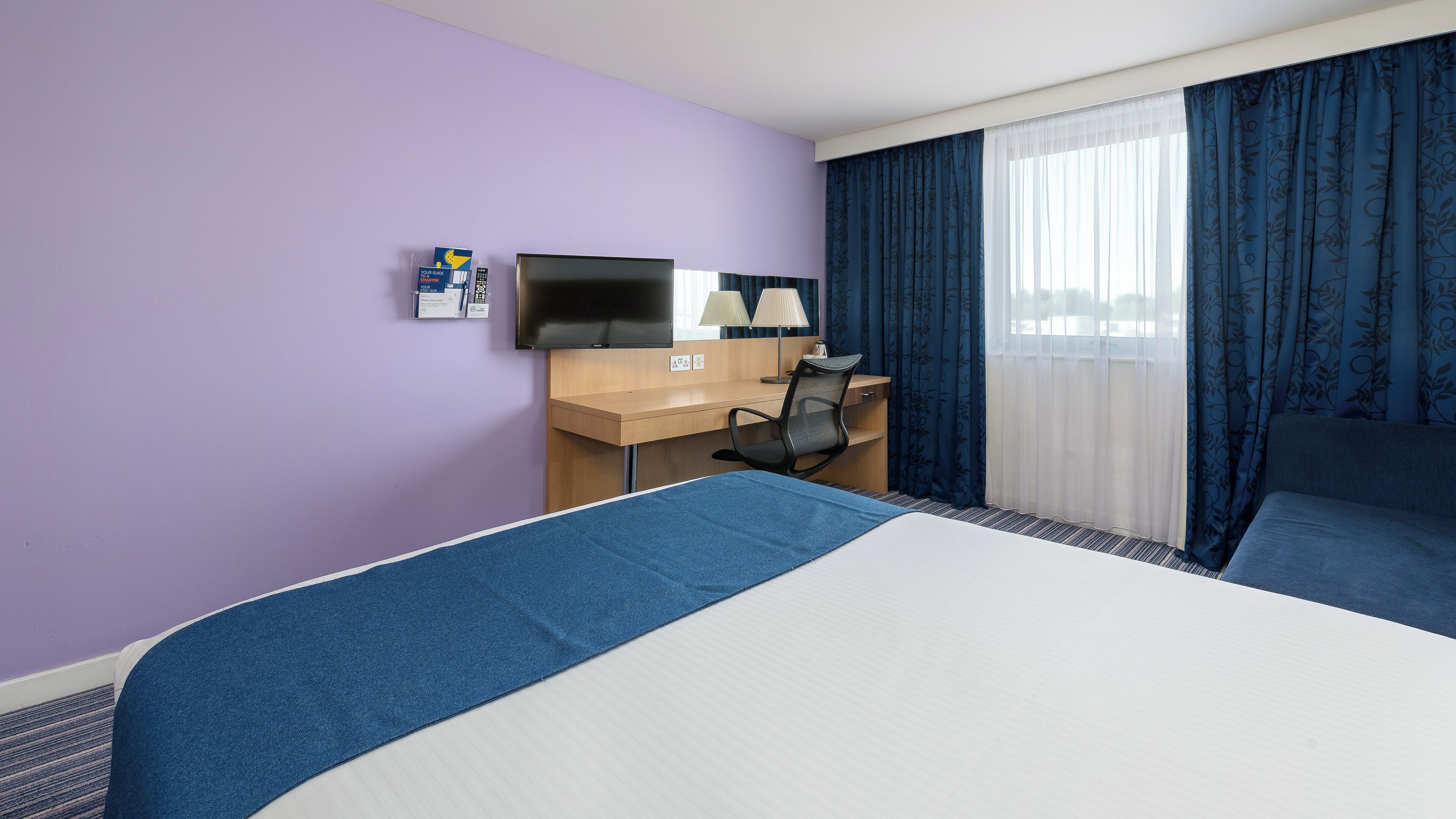 Una camera d'albergo con un letto, una scrivania con una sedia, una televisione e una finestra con tende.