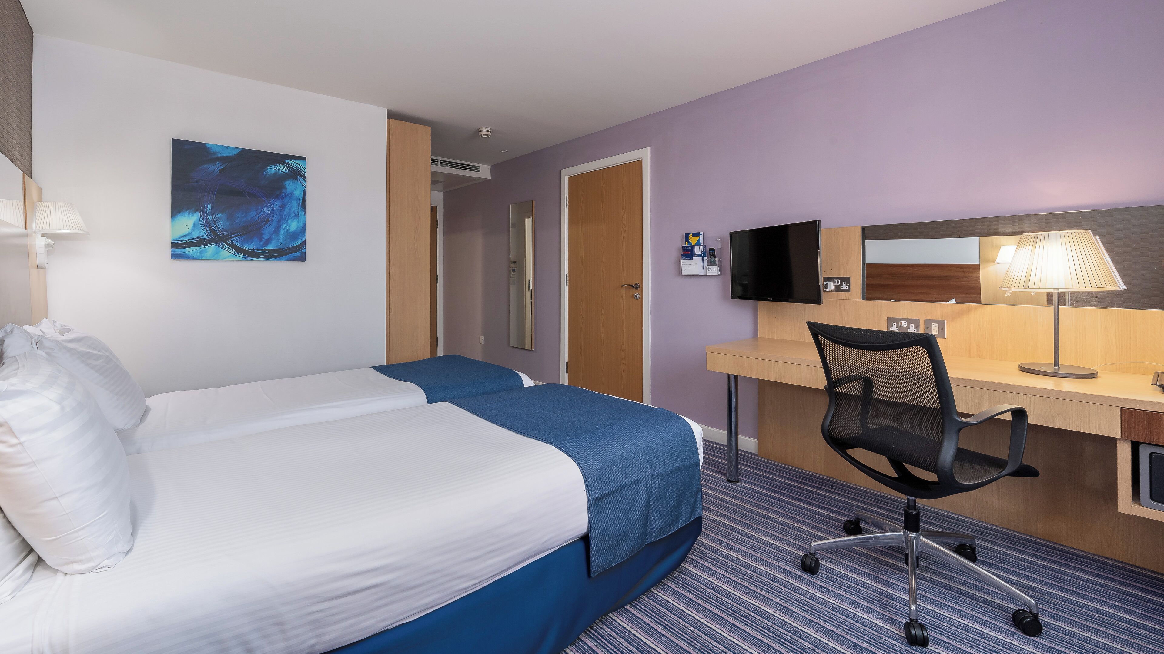 A hotel room with two beds, a desk, a chair, a TV, and a painting on the wall.
